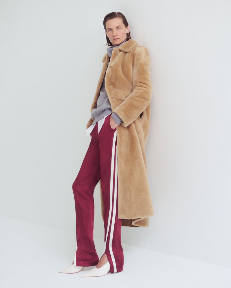 Woman in a beige coat with maroon pants and white shoes leans against a white wall in a full-length studio shot.