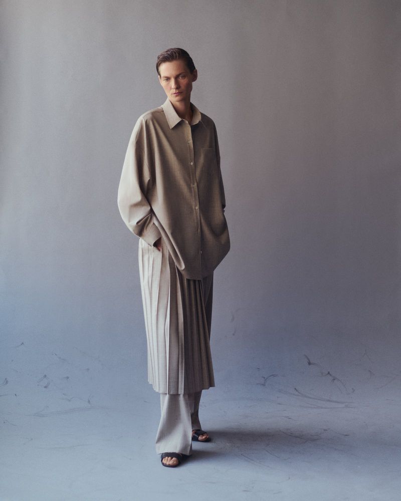 Full-length studio shot of a young adult woman in a tan, button-down shirt and pleated skirt with neutral background and soft, diffused lighting.