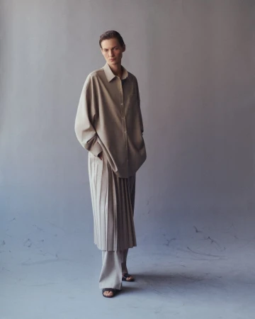 Full-length studio shot of a young adult woman in a tan, button-down shirt and pleated skirt with neutral background and soft, diffused lighting.