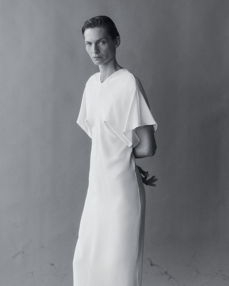 Elegant portrait of a woman in a white dress against a grey backdrop. The image is black and white, emphasizing textures and minimalist design.