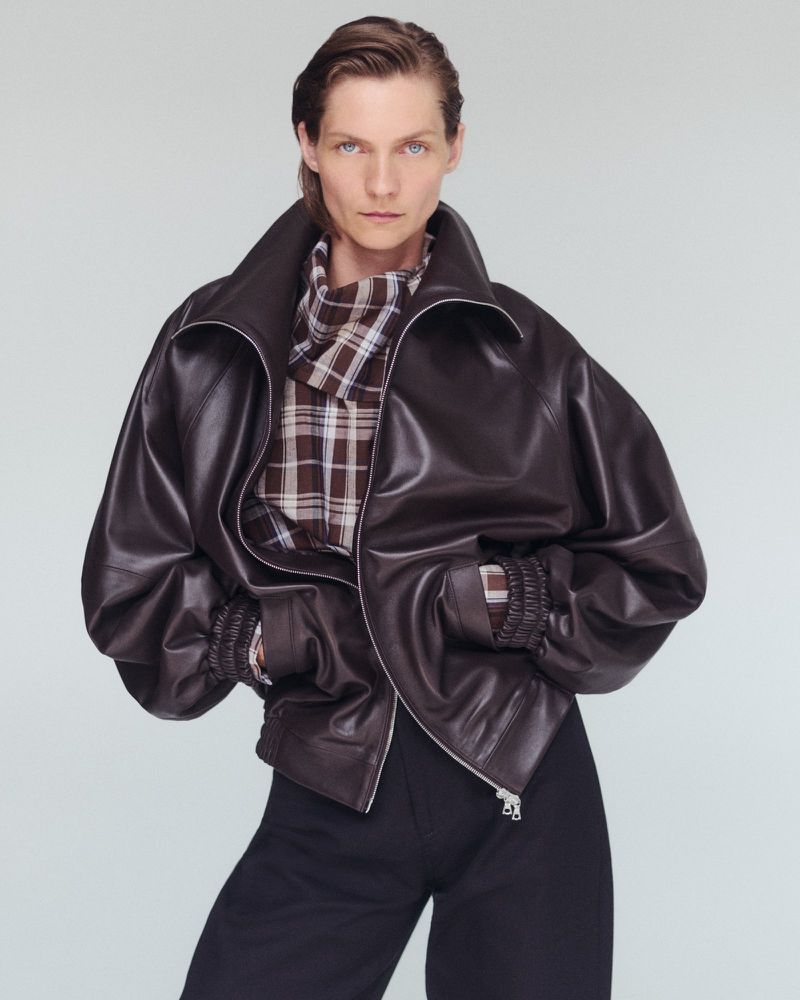 Woman in a dark brown leather bomber jacket with a plaid shirt underneath, standing against a light gray background in a studio.