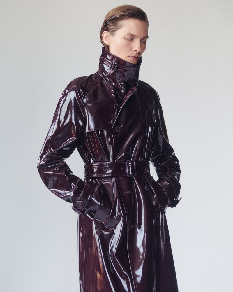 Adult woman in a shiny, dark red trench coat with a belt and high collar against a neutral background in a studio fashion shot.
