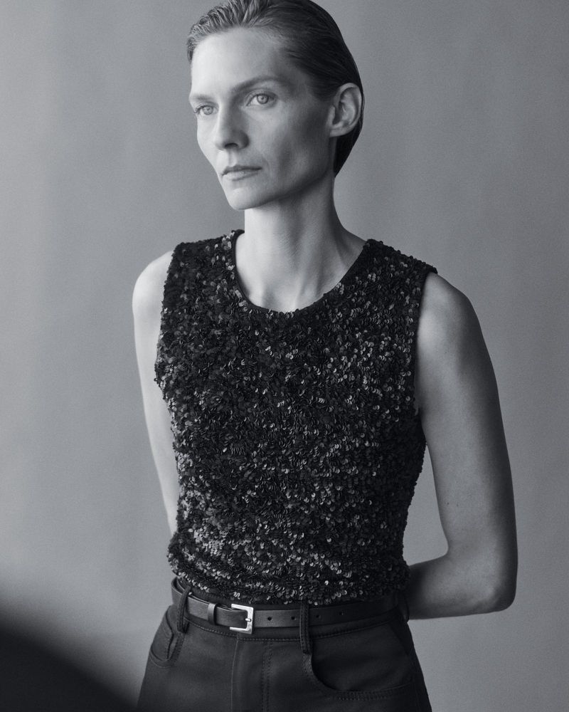Black and white portrait of a woman in a dark sequined top and dark pants looking off camera against a neutral background.