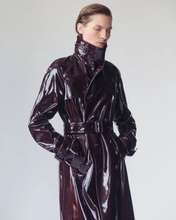 Model Karolin Wolter wears a glossy deep burgundy belted patent trench coat against a minimal studio background.