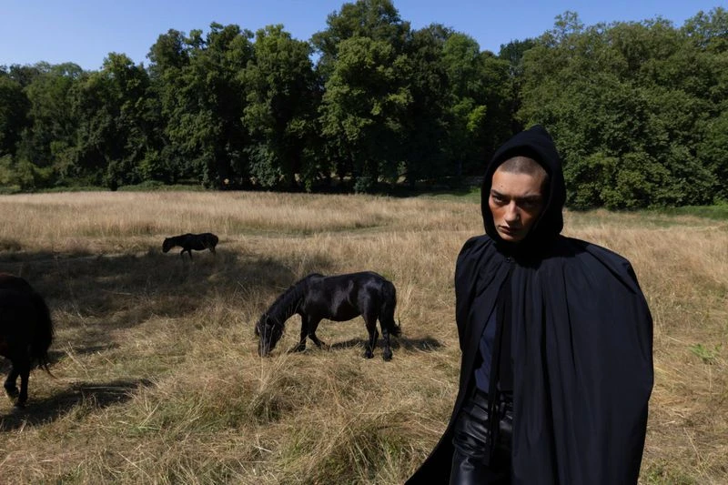 Model Isa Nilwik wearing a black hooded cloak in a grassy field with black horses for Mastermind Magazine.