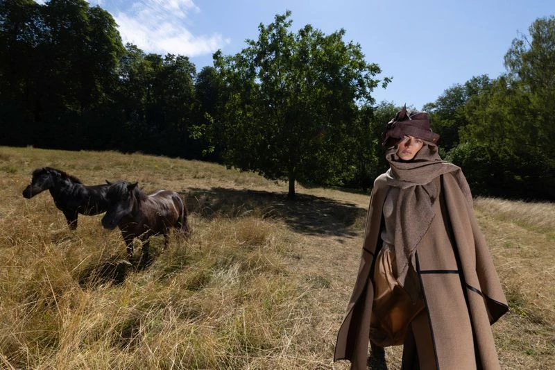 Model wearing brown cape and matching hat standing in a grassy field with two dark horses