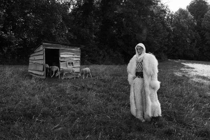 Model Isa Nilwik in a fluffy white fur coat and head covering standing in a grassy field near a wooden shed with sheep