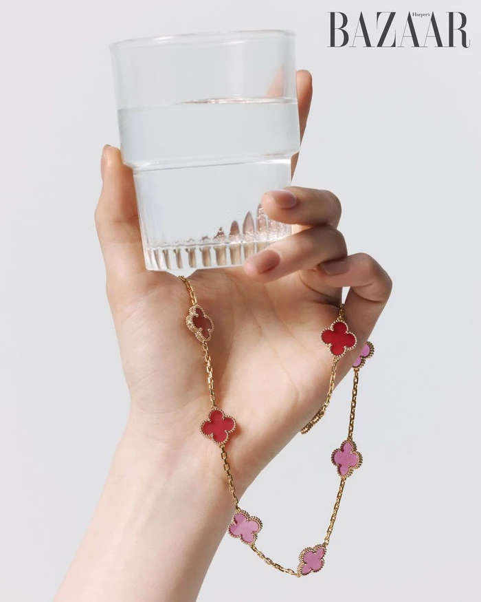 Close-up of a hand holding a glass of water and a gold clover necklace against a plain background