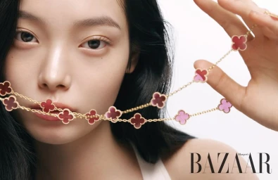 Close-up portrait of a woman holding a gold necklace with pink and red clover motifs near her face against a white background.