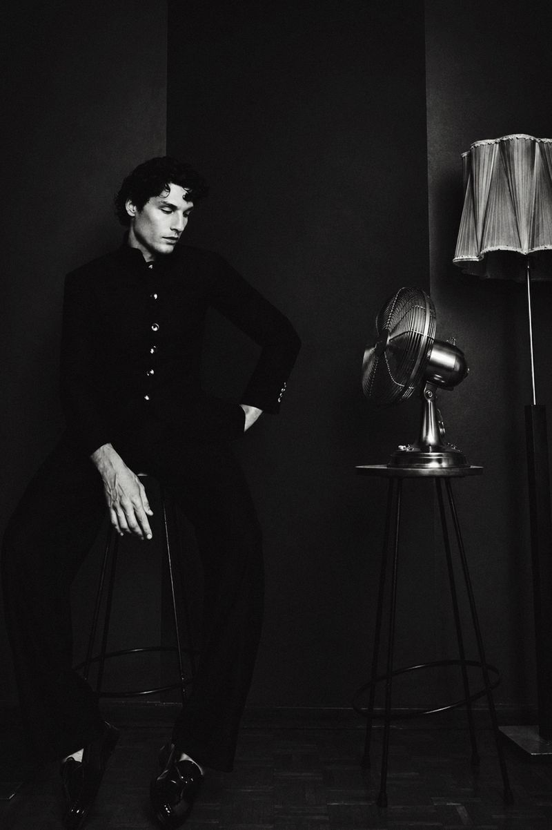 Monochrome portrait of a man seated on a stool next to a metallic desk fan and floor lamp in a dark room.