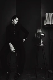 Monochrome portrait of a man seated on a stool next to a metallic desk fan and floor lamp in a dark room.