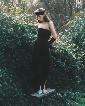 Model standing on a wooden plank wearing a black strapless dress, matching hat, gloves, and white boots in a lush wooded area.