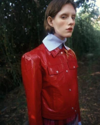 Model in red patent leather jacket and light blue collar shirt standing outdoors in a lush forest setting.