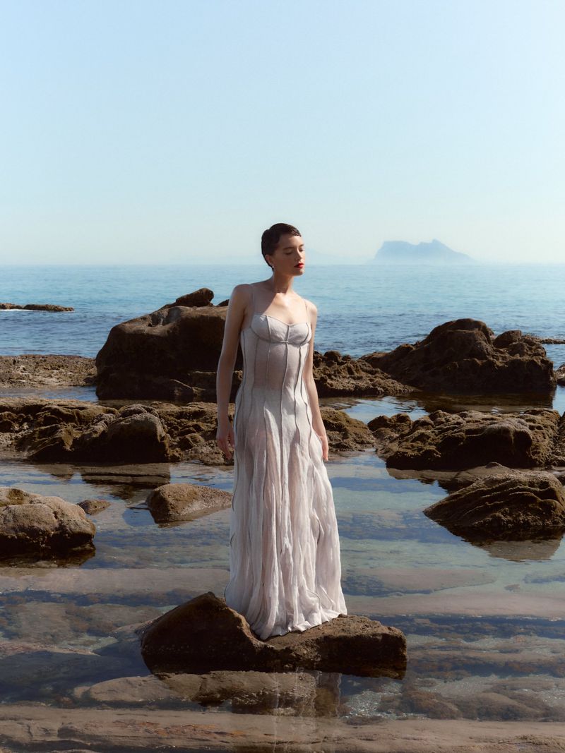 Model wearing a sheer long dress standing on a wet rock at the shoreline under clear daylight