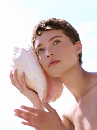 Model Loane Normand holding a large seashell to her ear against a bright sky for Harper's Bazaar Greece by Marina Testino