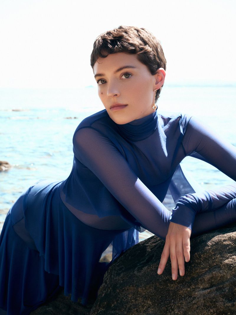 Model Loane Normand leaning over a dark rock by the sea in a blue sheer long sleeve top for Harper's Bazaar Greece.
