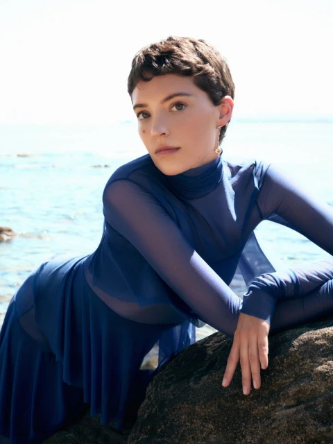 Model Loane Normand leaning over a dark rock by the sea in a blue sheer long sleeve top for Harper's Bazaar Greece.