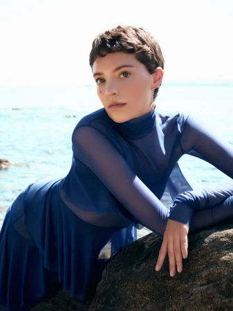 Model Loane Normand leaning over a dark rock by the sea in a blue sheer long sleeve top for Harper's Bazaar Greece.