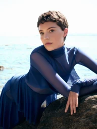 Model Loane Normand leaning over a dark rock by the sea in a blue sheer long sleeve top for Harper's Bazaar Greece.