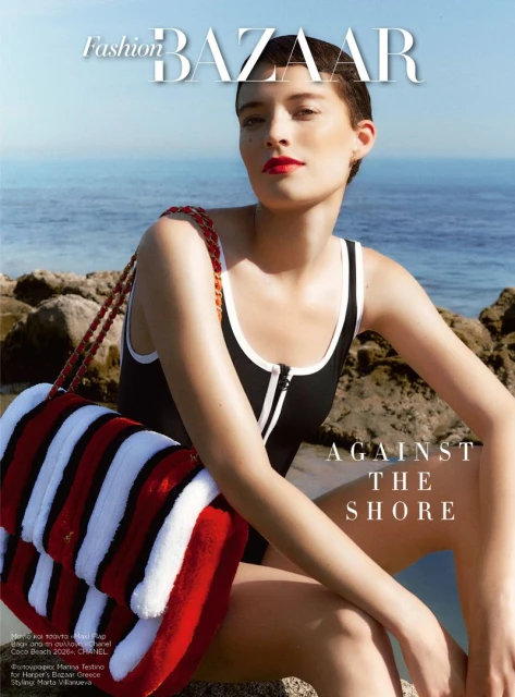 Model Loane Normand posing on coastal rocks with a large striped red and white bag for Harper's Bazaar Greece.