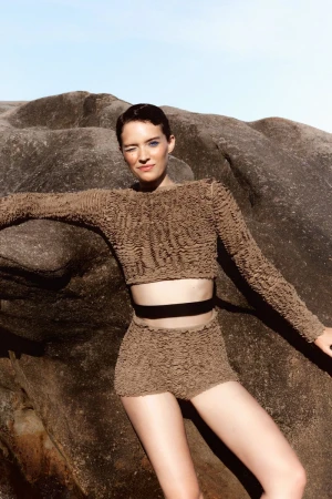 Model Loane Normand leaning against rock formations wearing textured brown top and shorts for Harper's Bazaar Greece.