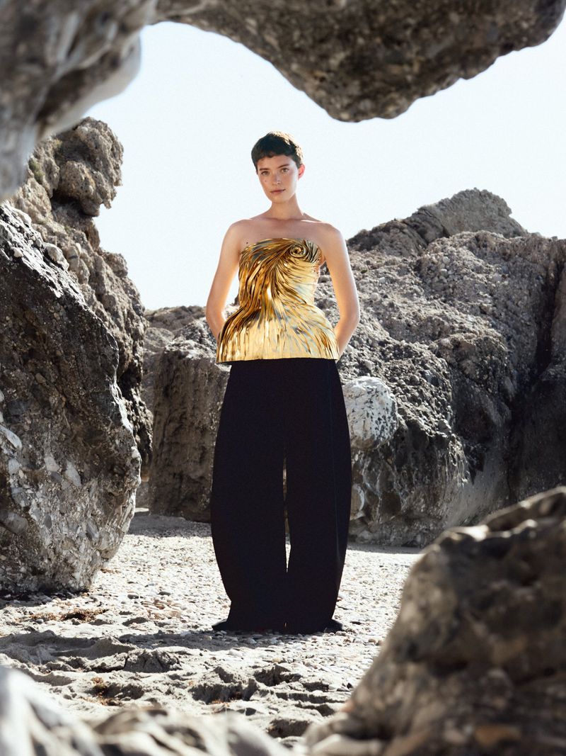 Model wearing a strapless metallic gold sculpted top and black trousers framed by rocky beach formations