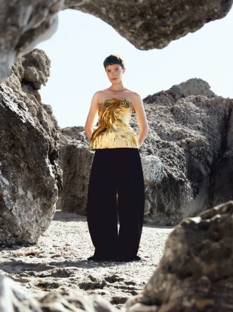 Model wearing a strapless metallic gold sculpted top and black trousers framed by rocky beach formations
