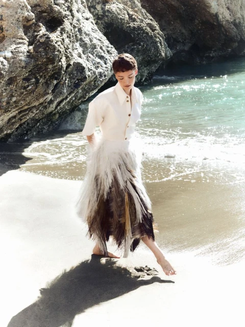 Model Loane Normand walking barefoot on a sandy beach wearing a white shirt and textured skirt for Harper's Bazaar Greece.