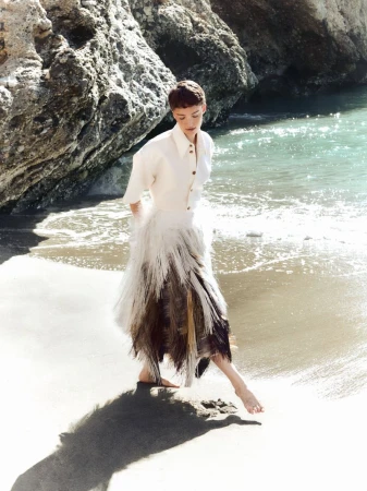 Model Loane Normand walking barefoot on a sandy beach wearing a white shirt and textured skirt for Harper's Bazaar Greece.