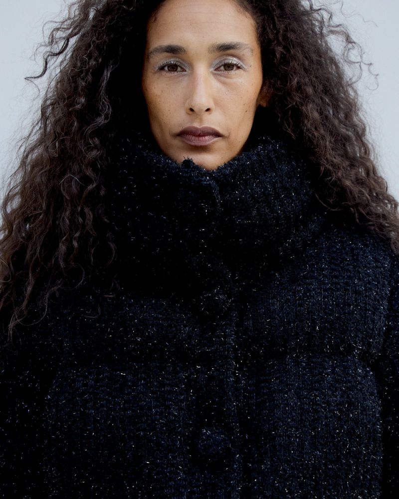Portrait of a woman with long dark curly hair wearing a textured tweed puffer jacket against a pale grey background.