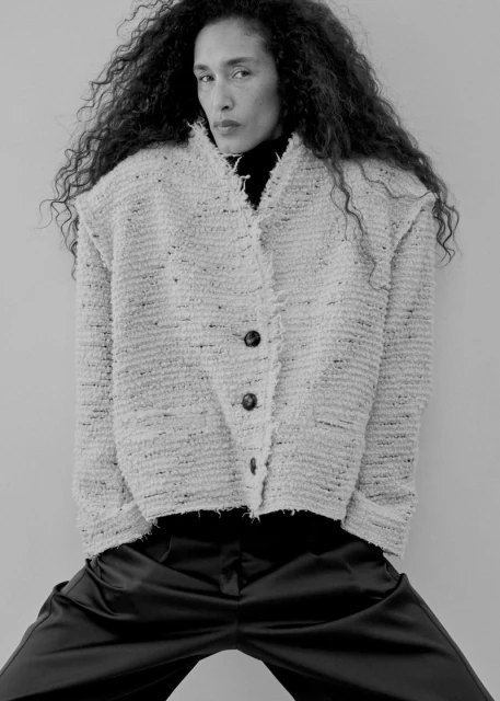 Model with long dark curly hair wearing a textured tweed jacket and dark trousers against a plain light background.
