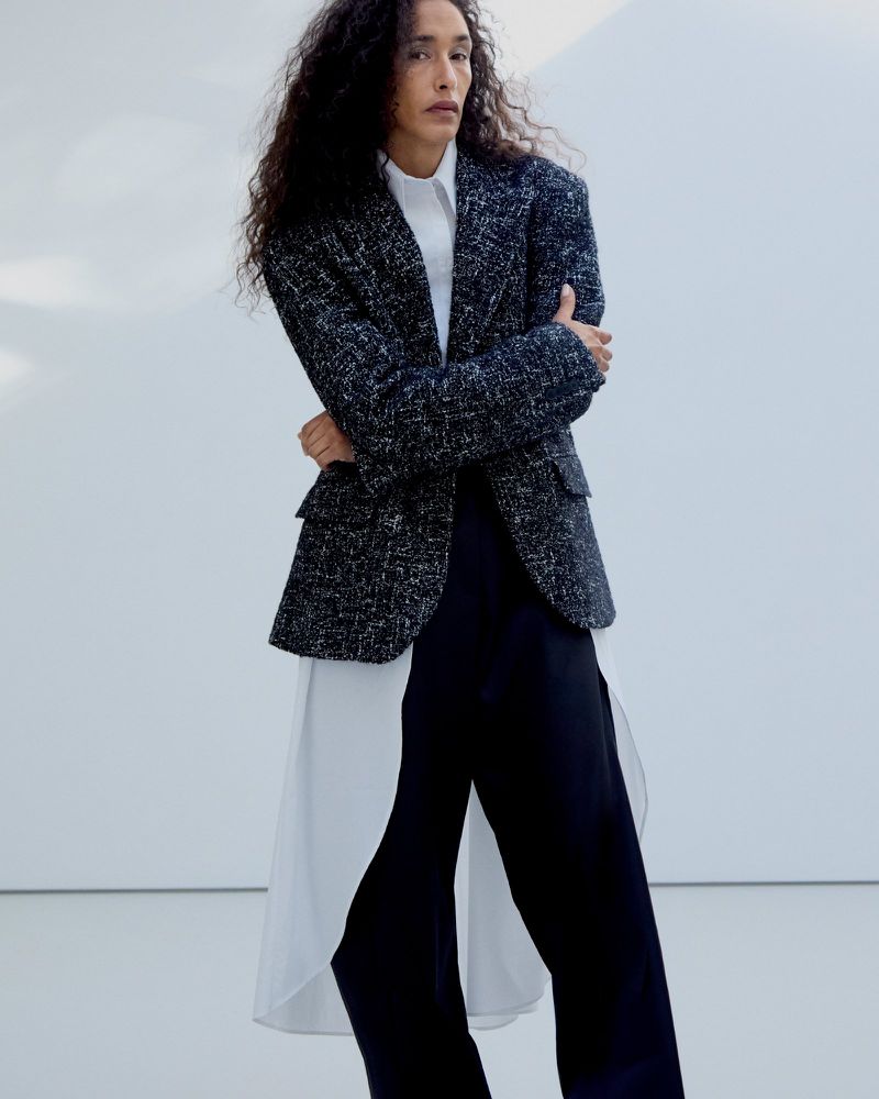 Model with long curly hair wearing a dark tweed blazer over a white shirt and dark trousers against a white backdrop.