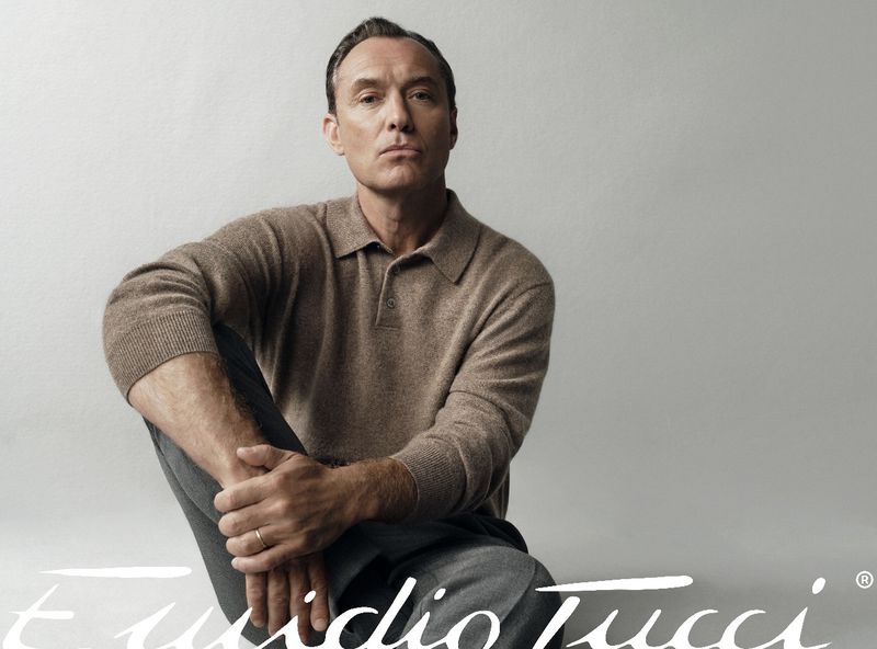 Jude Law seated wearing a brown polo shirt and dark trousers against a light grey studio background for El Corte Ingles.