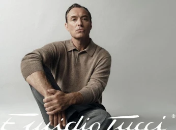 Jude Law seated wearing a brown polo shirt and dark trousers against a light grey studio background for El Corte Ingles.