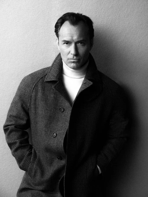 Portrait of Jude Law wearing a patterned coat and turtleneck sweater against a textured neutral background
