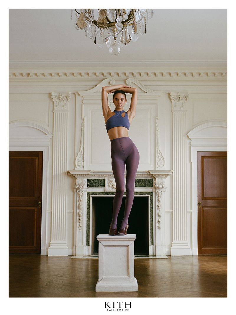 Model standing on a white pedestal in a luxury interior wearing activewear with arms raised above head