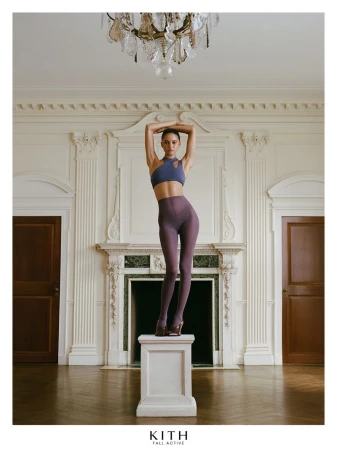 Model standing on a white pedestal in a luxury interior wearing activewear with arms raised above head