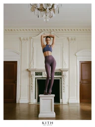 Model standing on a white pedestal in a luxury interior wearing activewear with arms raised above head