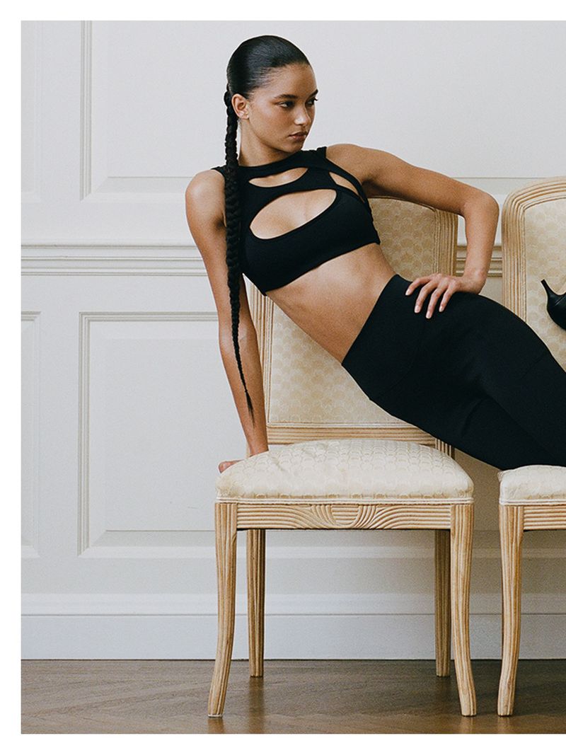 Model in a black cutout crop top leaning over ornate wooden chairs against white wainscoting