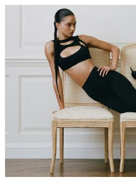 Model in a black cutout crop top leaning over ornate wooden chairs against white wainscoting