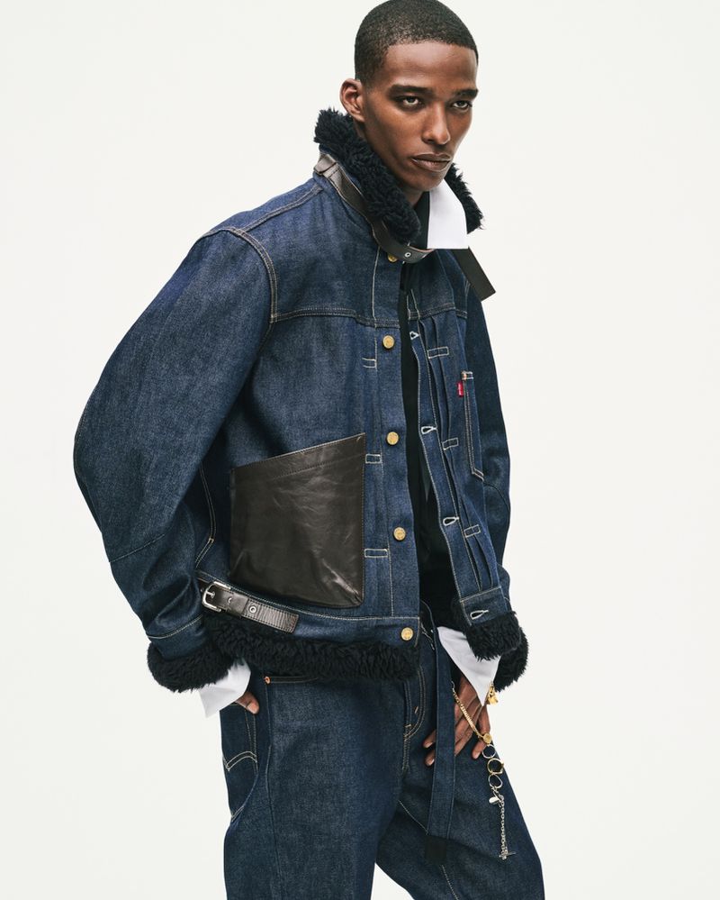 Model wearing dark denim jacket with shearling collar and leather pocket details against a white background
