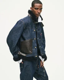Model wearing dark denim jacket with shearling collar and leather pocket details against a white background