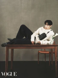 Man in white sweater and black trousers reclining on a wooden table wearing a crossbody bag for Vogue Hong Kong editorial