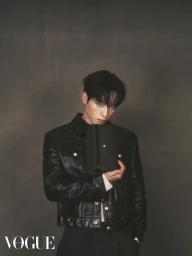 Man wearing a black leather jacket holding a small dark pouch against a textured dark grey backdrop for Vogue Hong Kong.