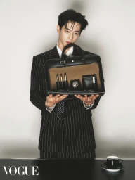 Man in a pinstripe suit holding open a black briefcase containing pens and a watch against a white background