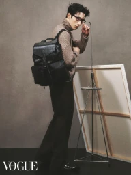 Man wearing glasses and beige turtleneck carrying a large black leather backpack in a studio with an artist easel