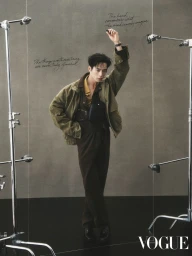 Man standing in an olive green jacket and brown trousers posed between studio light stands against a textured grey backdrop.