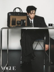 Man wearing a black pinstripe suit sitting at a metal table with an open leather briefcase and coffee cup against a white background