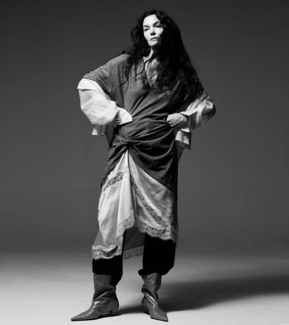 Full-length black and white editorial portrait of a woman wearing a layered dress and boots against a seamless studio backdrop.