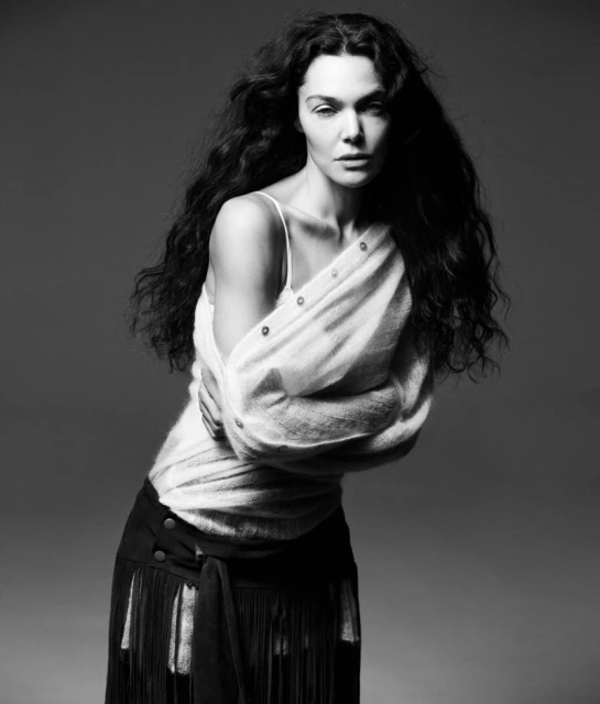 Monochrome portrait of a woman with long dark wavy hair wearing an off-shoulder white top and dark skirt against a grey background.